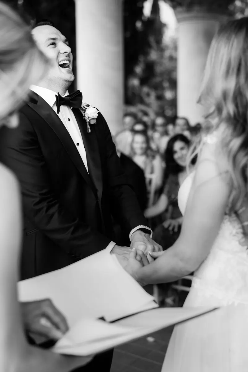 Black and white photo of the groom laughing as he reads his vows